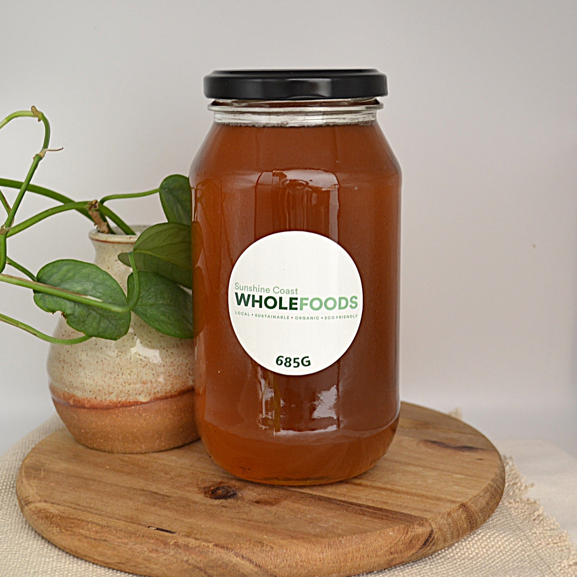 685g Jar of Sunshine coast honey with a Whole Foods label on a wooden board, next to a plant.